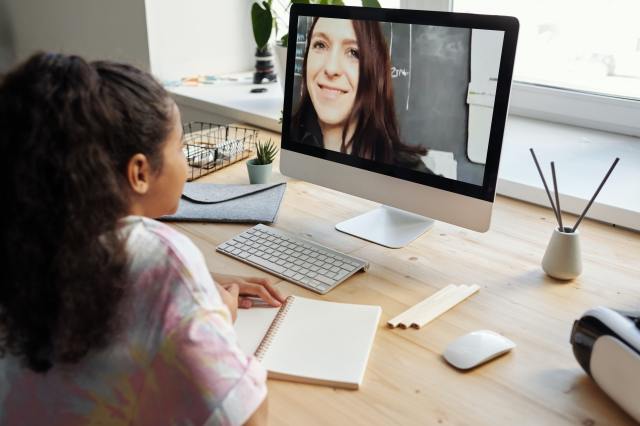 Girl at a desk watches her teacher on a computer monitor
